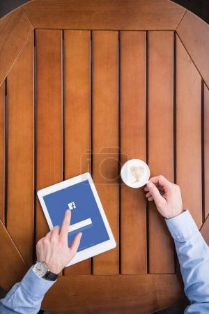 cropped image of man sitting with coffee and loaded facebook page on tablet 