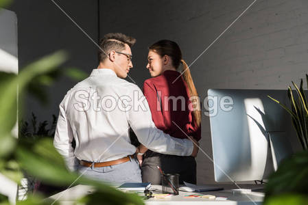 rear view of managers embracing at workplace, office romance concept