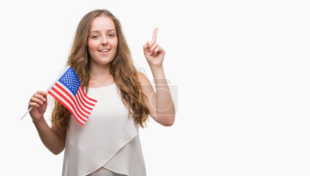 Young blonde woman holding flag of USA surprised with an idea or question pointing finger with happy face, number one