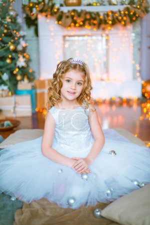 Cute little girl with curly blond hair at home near a Christmas tree with gifts and garlands and a decorated fireplace sitting on plaids and pillows