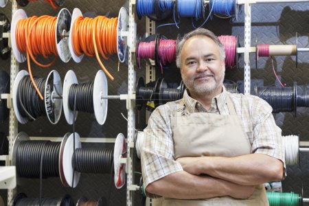 Portrait of a happy mature salesperson standing in front of electrical wire spool with arms crossed in hardware store