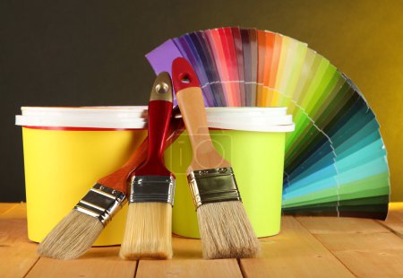 Paint pots, paintbrushes and coloured swatches on wooden table on dark yellow background