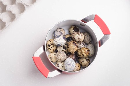 Quail eggs in a red saucepan, cardboard packaging on white stone table