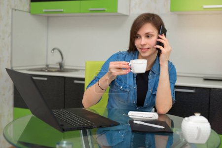 beautiful young girl sitting in the kitchen at the table with a laptop drinking tea and talking on the phone