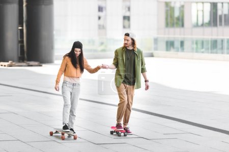 brunette woman holding hands with man, riding on skateboard in city