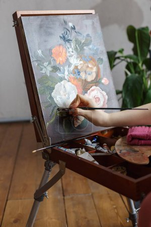 Close up on artist hands with brush painting flowers picture on an easel. Art, creativity, hobby concept.