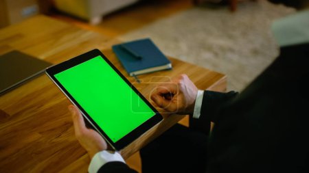 At Home Man Sits at His Desk and Holds Tablet Computer with Green Screen on it. His Apartment is Done in Yellow colours and is Warm.