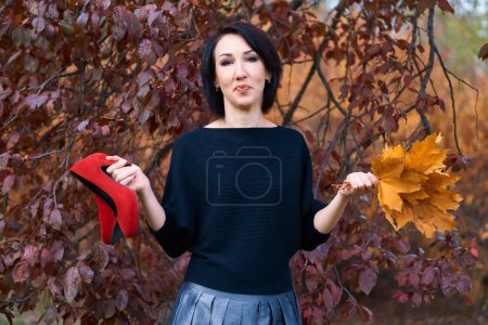 Beautiful elegant woman standing and posing with bouquet of yellow leaves and red shoes in autumn city park