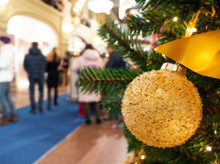 Golden ball, toy for Christmas tree in mall. Traditional decoration for New Year celebration. Interior of store with people on background.