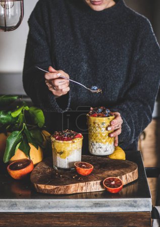 Young woman eating overnight oat bircher muesli with mango smoothie, buckwheat granola and fresh ripe berries from glass with spoon over grey concrete kitchen counter. Healthy, clean eating concept
