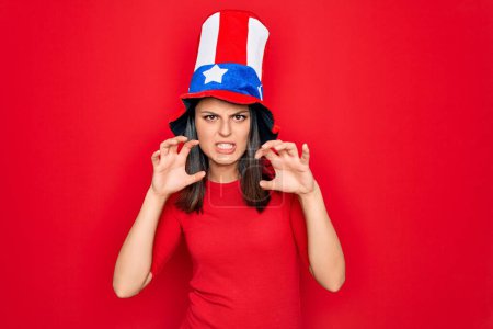 Young beautiful brunette woman wearing united states hat celebrating independence day smiling funny doing claw gesture as cat, aggressive and sexy expression