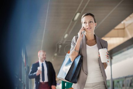 Portrait of young attractive businesswoman talking on smartphone and holding cup of coffee while walking against colleague