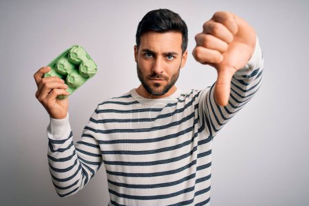 Young handsome man with beard holding carton box of fresh eggs over white background with angry face, negative sign showing dislike with thumbs down, rejection concept