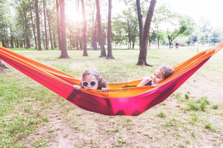 Happy little girls relax on hammock in nature. Children with sunglasses enjoy beautiful summer day in forest.