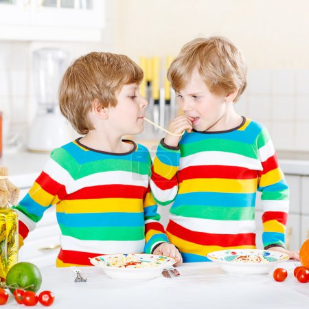 Two little kid boys eating spaghetti in domestic kitchen.